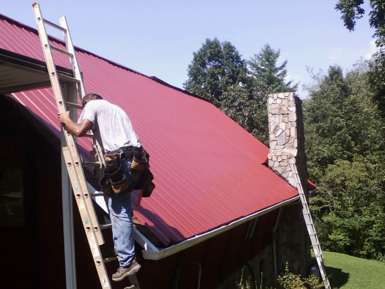 Roofer on a ladder beside a red metal roof and stone chimney — Soffit & Fascia Repair by Roofing Brothers in Calhoun City